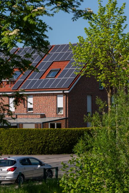 A modern brick house with solar panels on the roof, surrounded by lush greenery, under a clear blue sky.