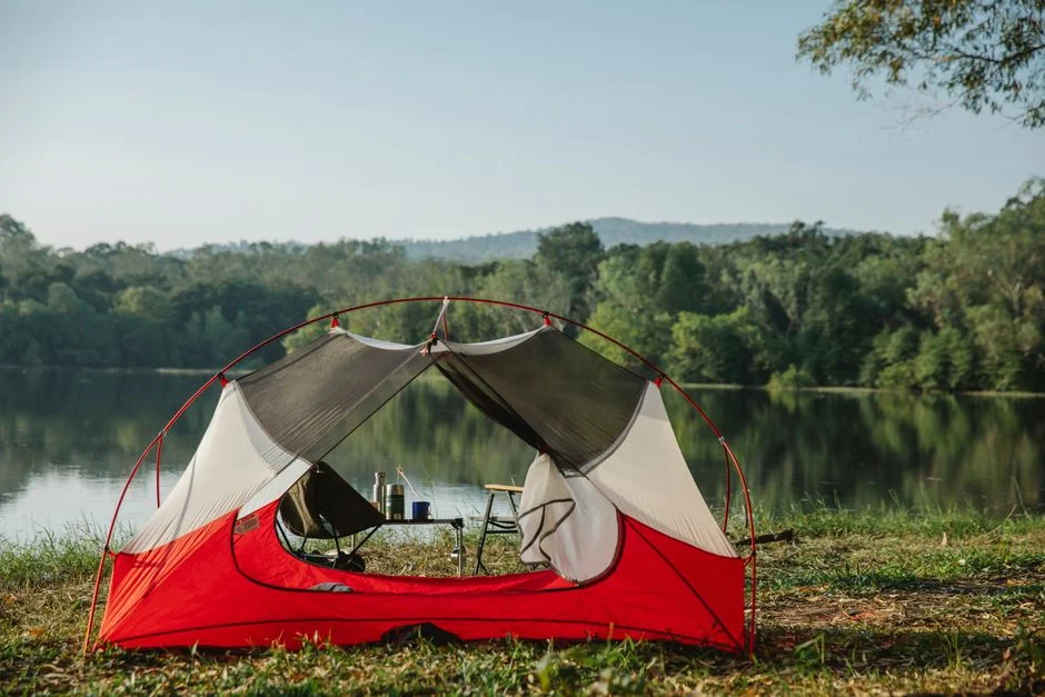 Tent with folding chairs and table on coast against trees reflecting in river under light sky