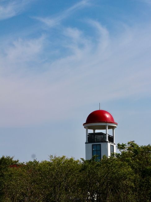 Lighthouse with red dome and blue sky backdrop, surrounded by lush greenery.