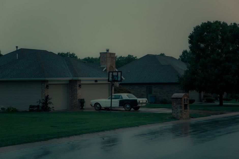 A classic car parked outside a suburban house near a basketball hoop on a wet road.