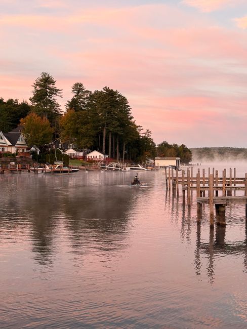 A tranquil lakeside scene captured at sunrise with mist over the water and pastel skies, offering a peaceful morning ambiance.