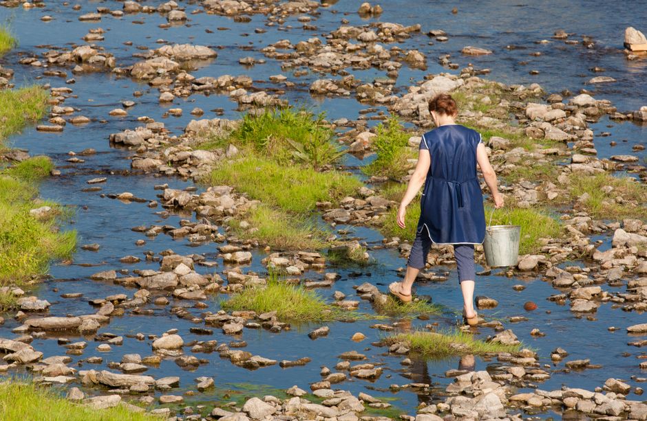 Woman crossing a shallow rocky river with a metal bucket in summer.