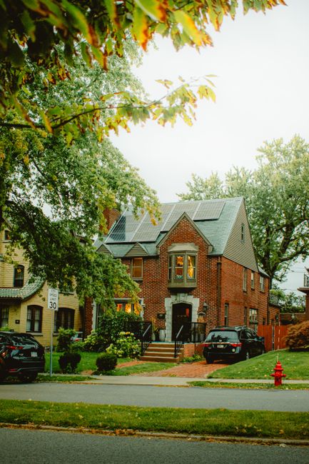 A picturesque brick house with solar panels, lush greenery, and fall foliage in a suburban neighborhood.