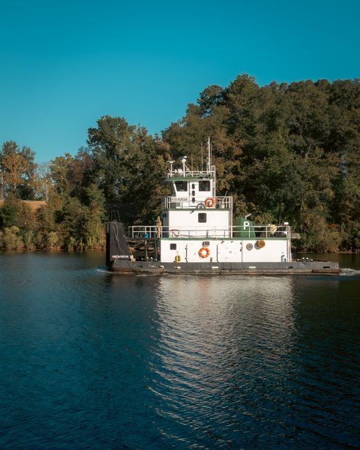A tugboat glides along a peaceful river surrounded by lush trees in Tuscaloosa, Alabama.
